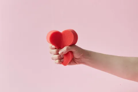 A hand squeezing a red foam heart against a pink background