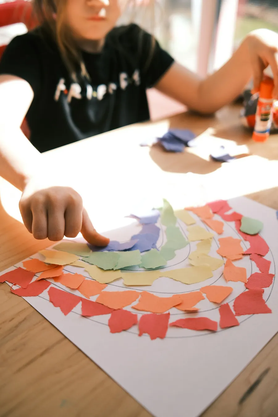 A child's finger pressing torn colored paper into a rainbow collage