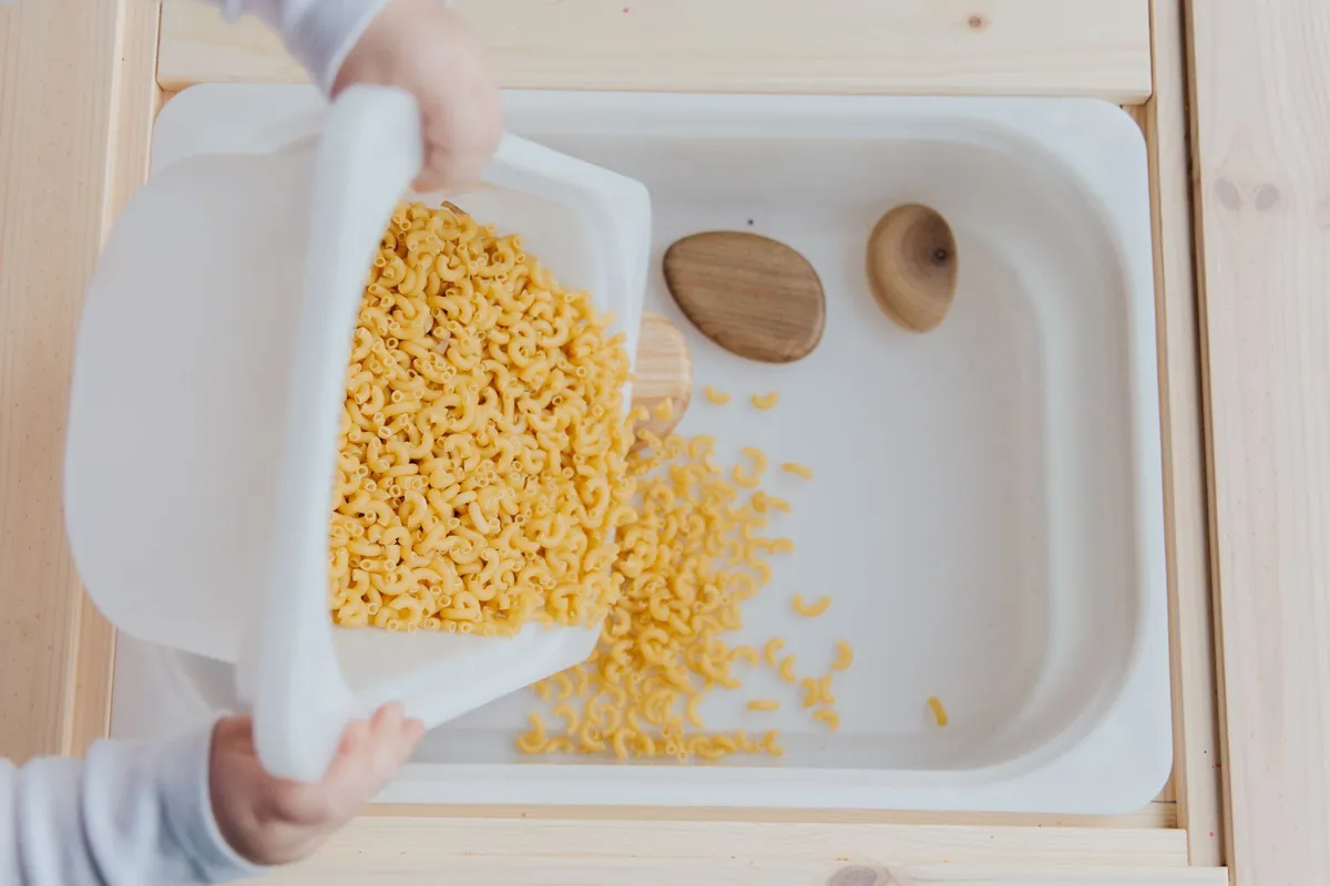 A child's hands putting dry pasta into white containers for sensory play