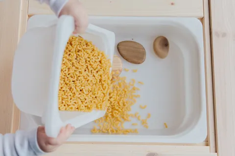 A child's hands putting dry pasta into white containers for sensory play